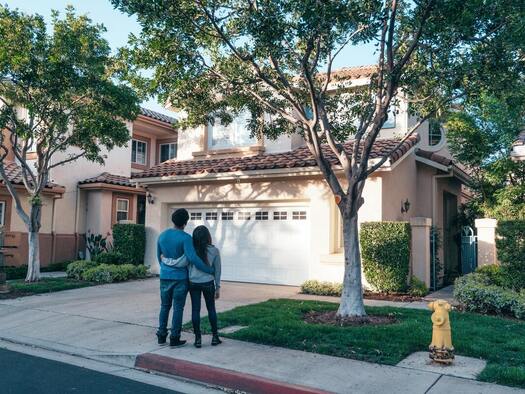 A couple holding each others waist standing on sidewalk in front of a house and admiring the house
